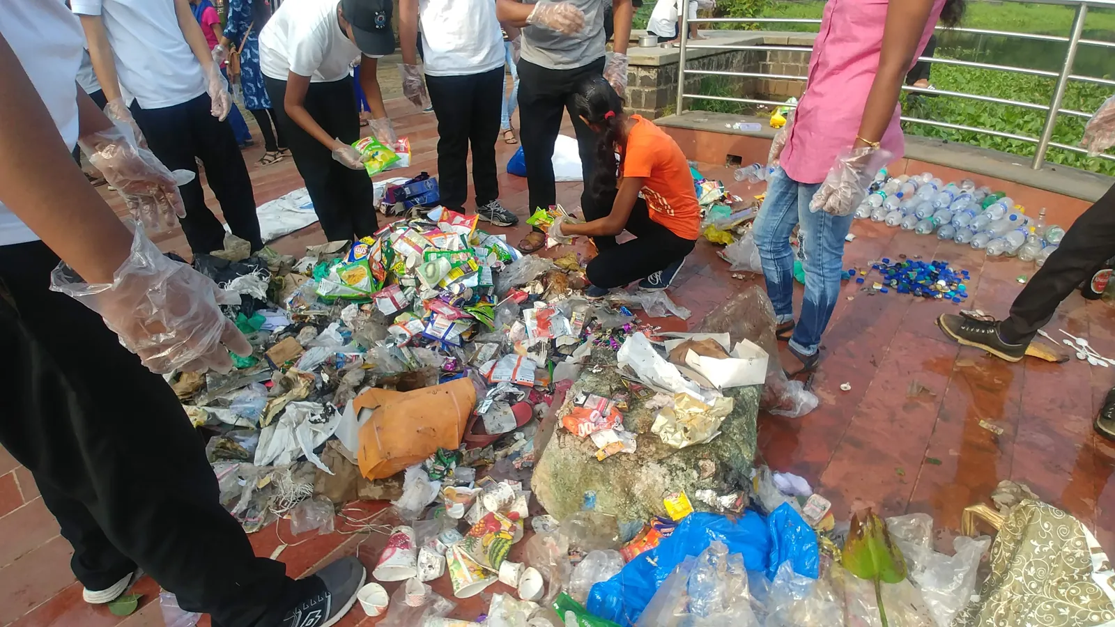 Gloved volunteers crouching around a large heap of mixed plastic packets, wrappers and bottles, sorting it by hand on a red-tiled lakeside walkway