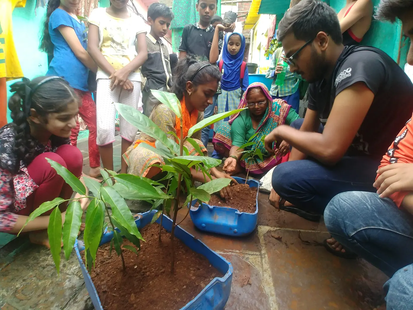 Women residents and Earth5R volunteers planting young saplings in blue crates of compost in a crowded lane of a Mumbai informal settlement