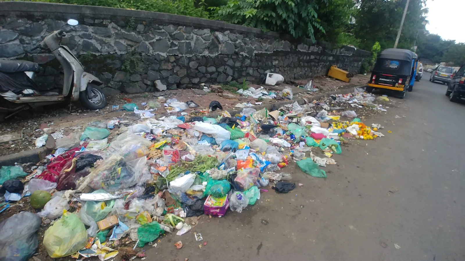 A long strip of mixed plastic bags, wrappers, flowers and paper litter along a lakeside road edge in Mumbai, an auto-rickshaw parked beyond
