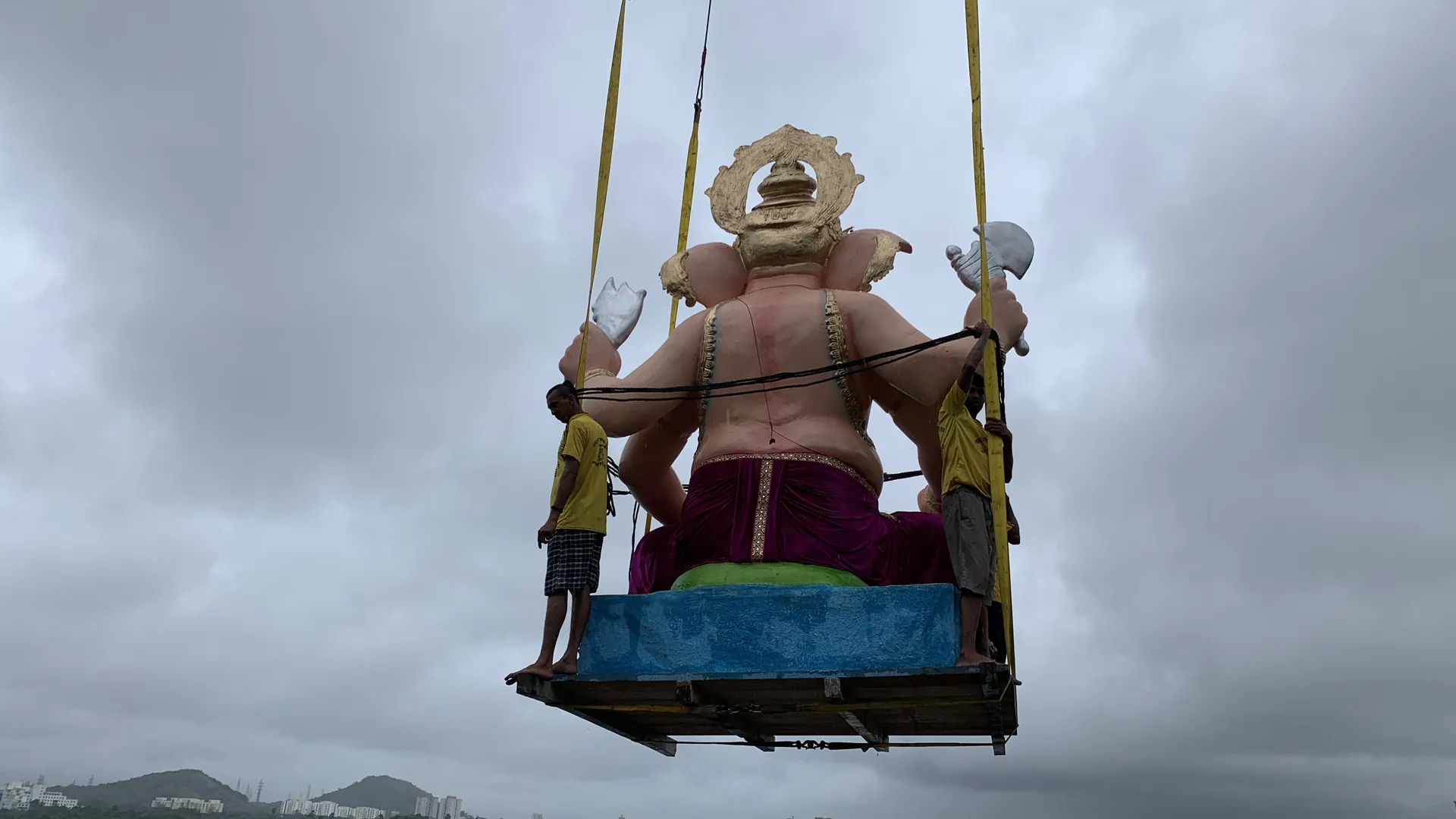A tall Ganesh idol on a crane platform, held by two men in yellow T-shirts, lifted against a grey monsoon sky above a lake shore on immersion day
