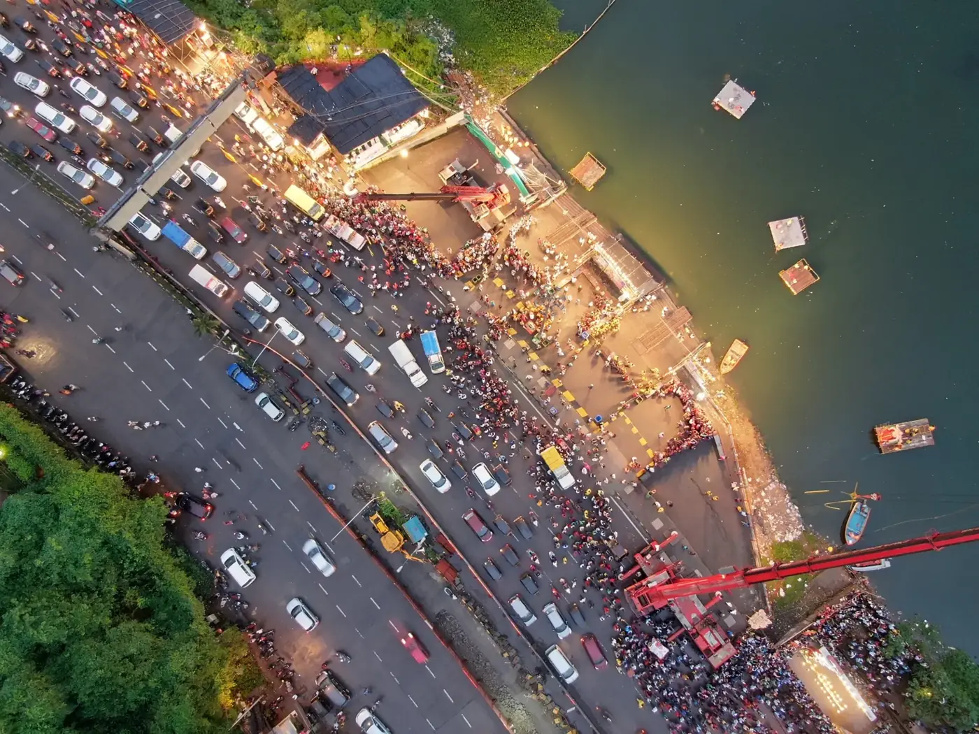 Aerial view at dusk of a lakeside immersion point packed with crowds and vehicles, a red crane reaching over the water and floating debris near the shore