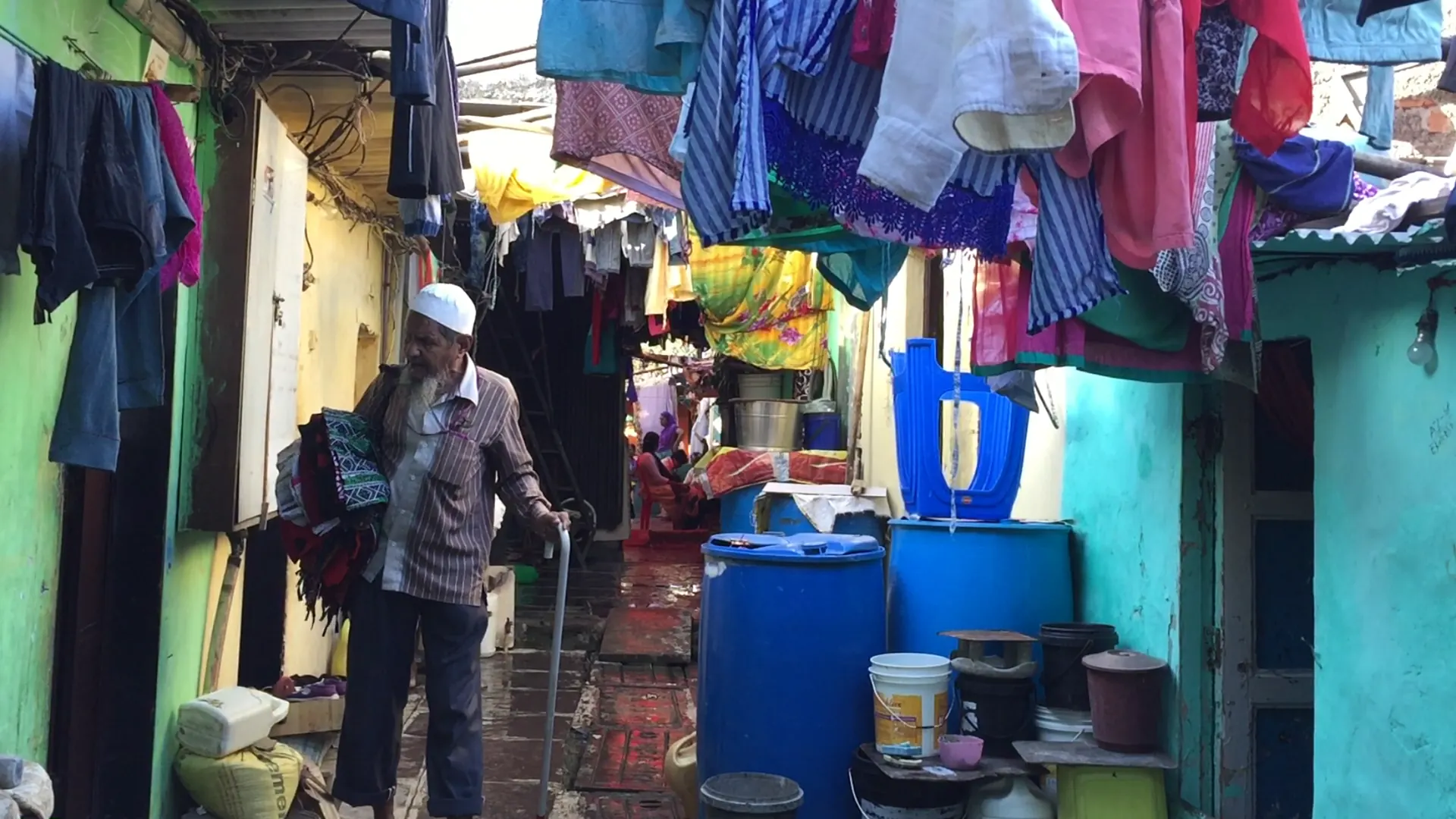 Blue plastic water drums and buckets lined up along a narrow residential lane, with an older man walking past laundry hung overhead