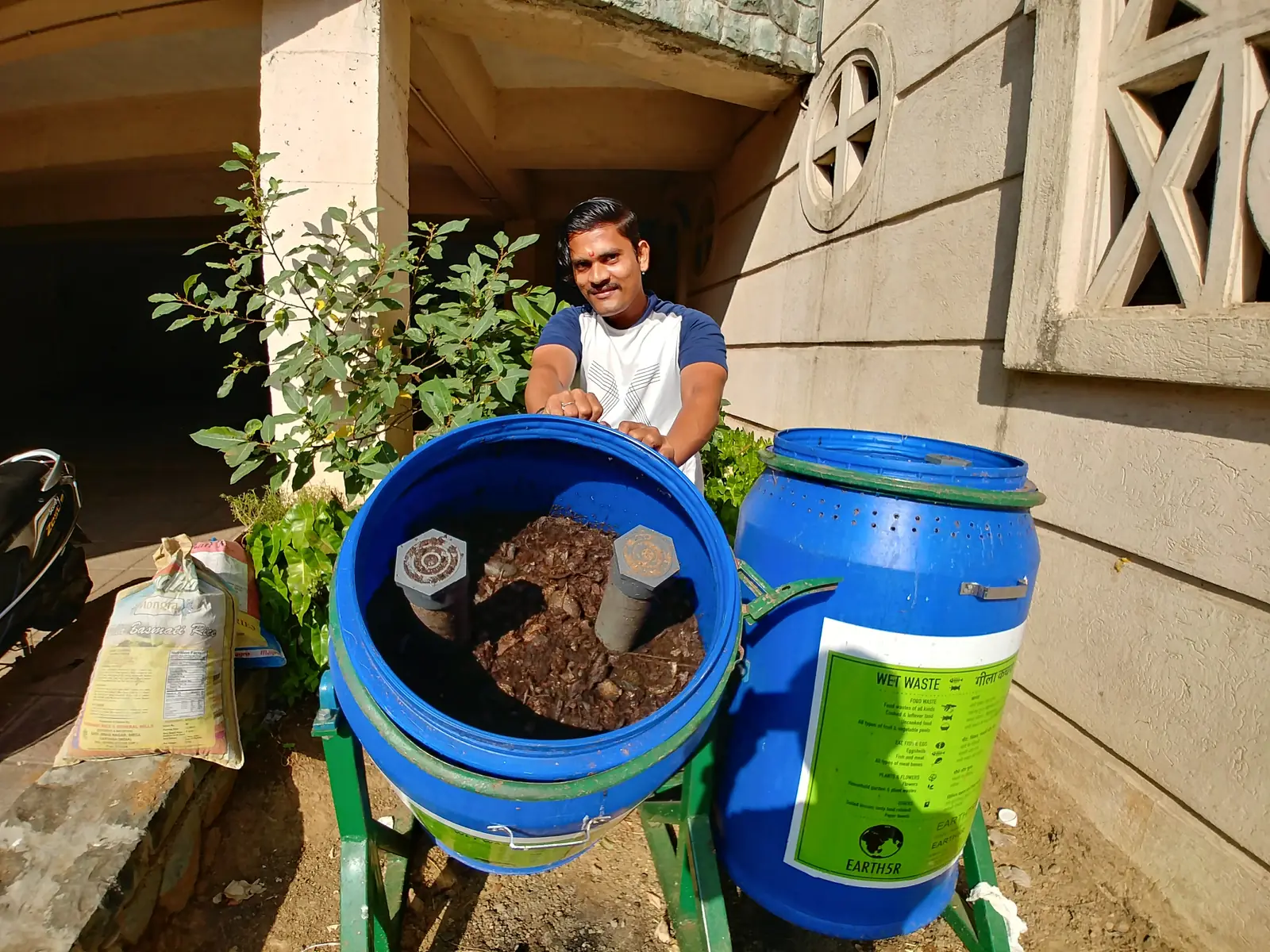 High school nutrition internship field work. Resident with blue and green home composting drums on a terrace, plants behind