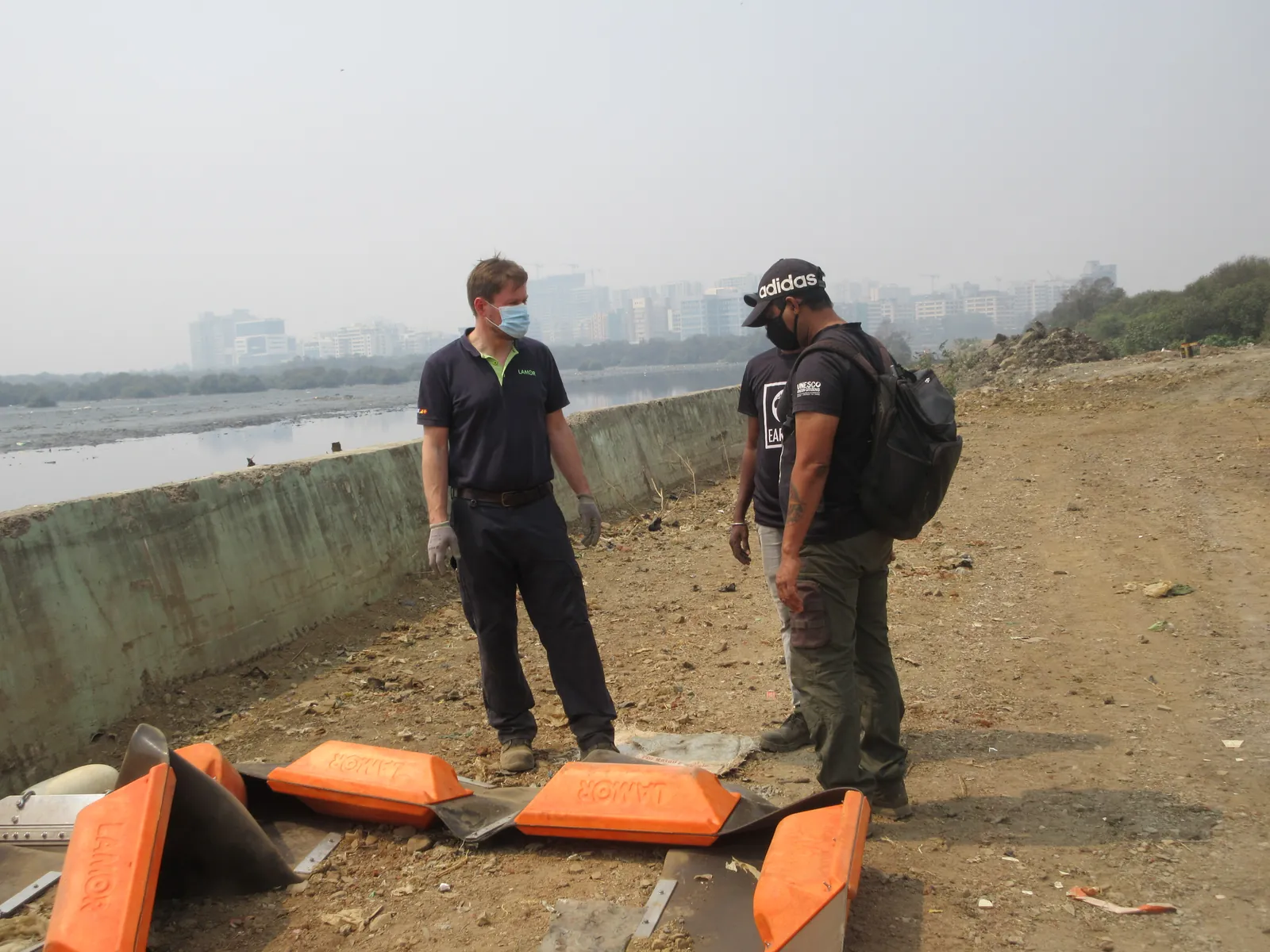 High school mechanical engineering internship field work. Two men with orange float sections of a trash boom on a dry riverbank