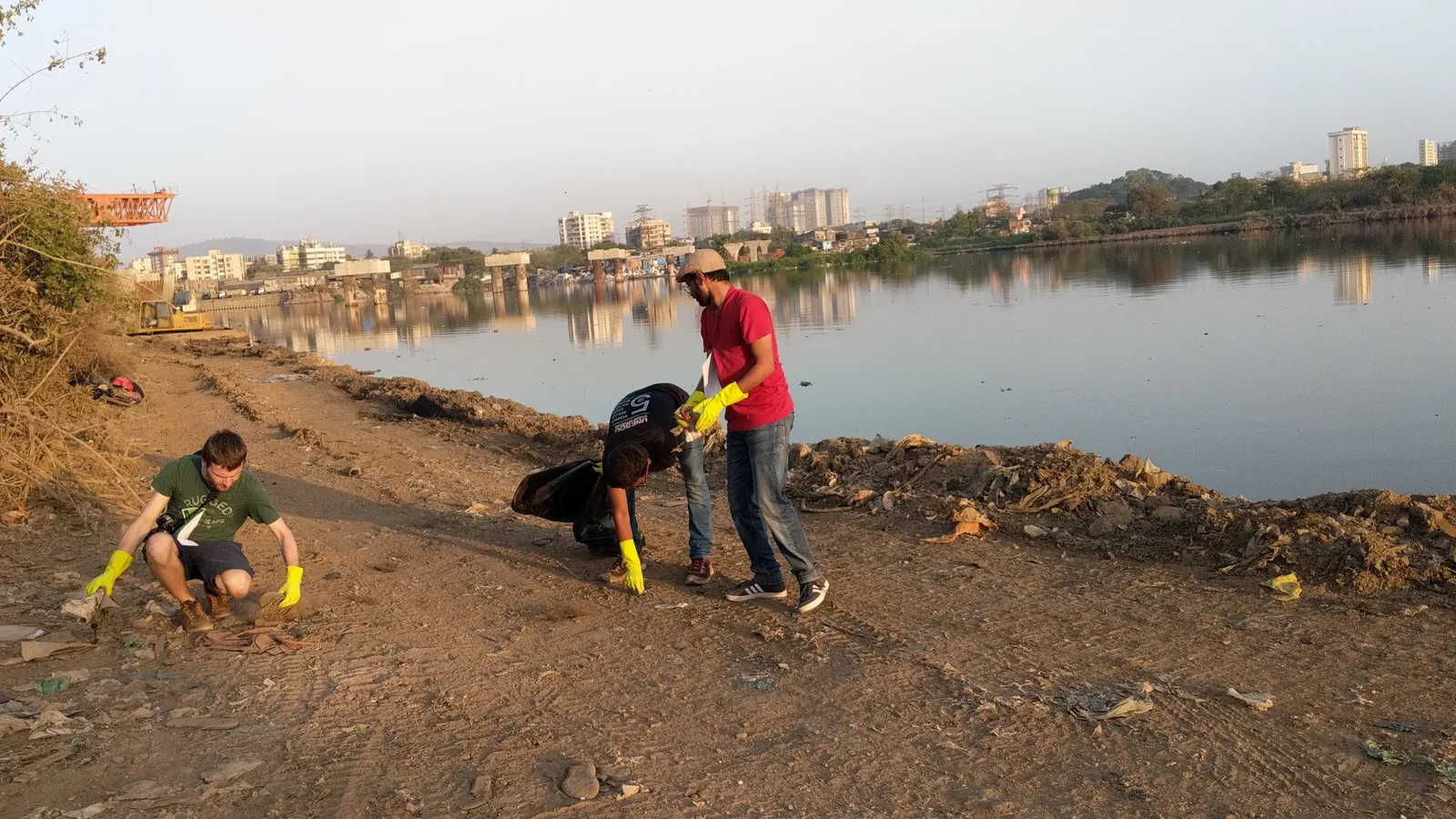 Geography internship field work. Earth5R volunteers picking waste on a muddy lake shore with the city skyline behind in Mumbai