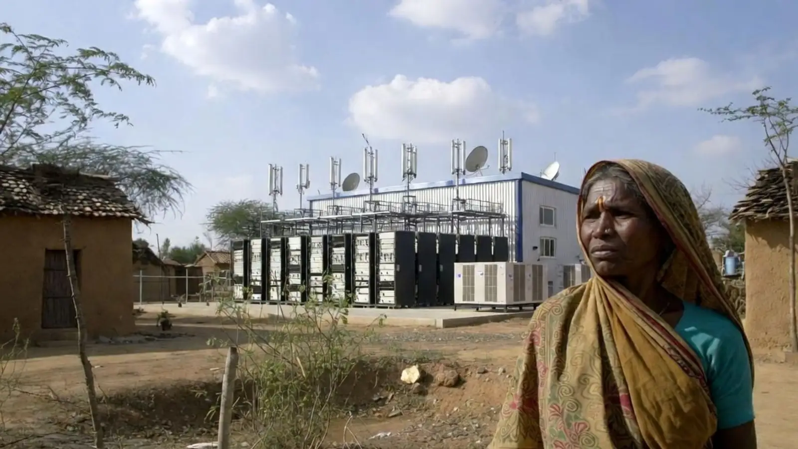 Rural woman in a sari standing in a dry, water-scarce village with mud houses, with a small data facility and cooling units behind her