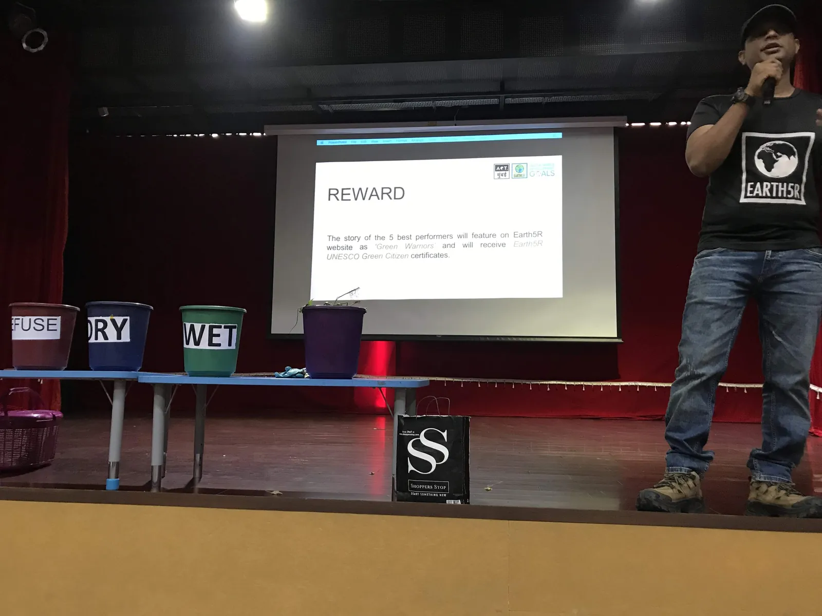High school social work internship field work. Earth5R speaker presenting on a school stage beside colour-coded waste bins