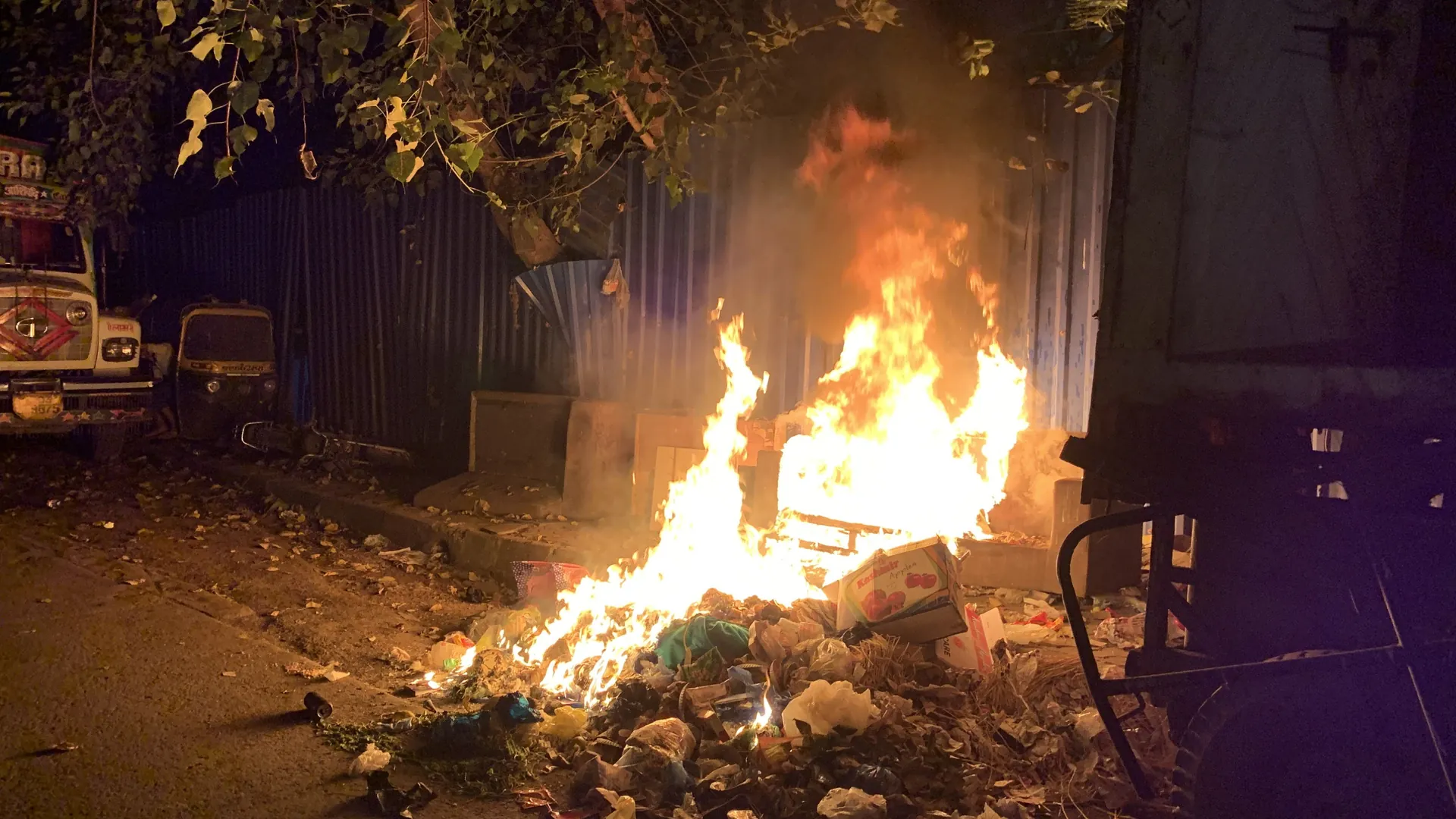 A heap of mixed waste and cardboard burning in a tall open fire at night beside a metal fence and parked trucks in Mumbai, smoke rising into the trees